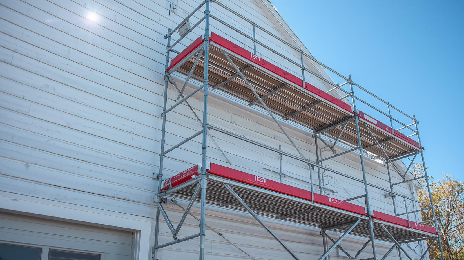 Bildet_viser_et_rammestillas_montert_langs_fasaden_pa_en_hvit_trebygning_med_flere_vinduer._Stillaset_bestar_av_aluminiumsrammer_horisontale_gangbaner_og_rode_plattformer_som_danner_a
